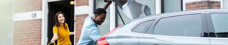 Man en vrouw bij huis laden auto in terwijl vrouw een grote hond uitlaat.